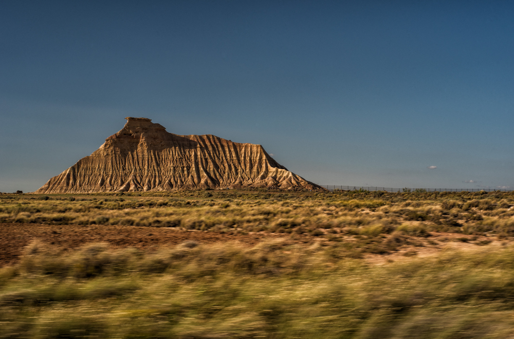 mountain looms over Bardenas Reales' semi-desert landscape. This photo captures a large, solitary butte or mesa rising from a flat desert plain. The formation has steep, striated sides with visible layers of rock, suggesting years of erosion. The foreground shows sparse desert vegetation, mostly dry grasses. The sky is a clear, deep blue, contrasting with the warm oranges and browns of the landscape.