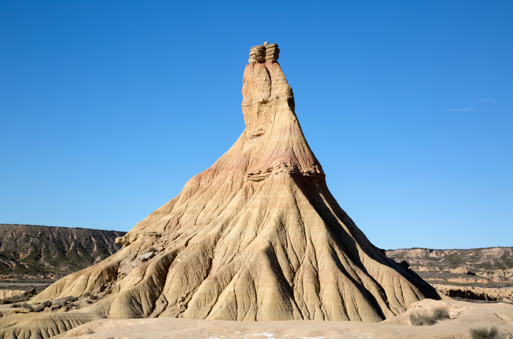 Iconic pinnacle rock formation rises from Bardenas Reales' arid plains. This image features a dramatic rock formation known as a hoodoo. It's a tall, conical spire of rock with a distinctive shape, broader at the base and tapering towards the top, with a small cap rock. The rock is light beige in color and shows clear signs of erosion with striations along its sides. It stands alone against a bright blue sky, dominating the landscape.