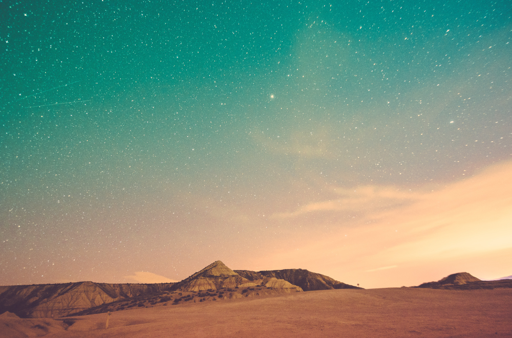 Night sky stretches over Bardenas Reales' desert landscape. This image captures a nighttime desert scene. In the foreground, there are sandy dunes or rocky formations. The sky is the most striking element, filled with countless stars creating a breathtaking display. The colors transition from a warm orange glow on the horizon to a deep teal in the upper sky. It gives a sense of the vastness and beauty of the desert at night.