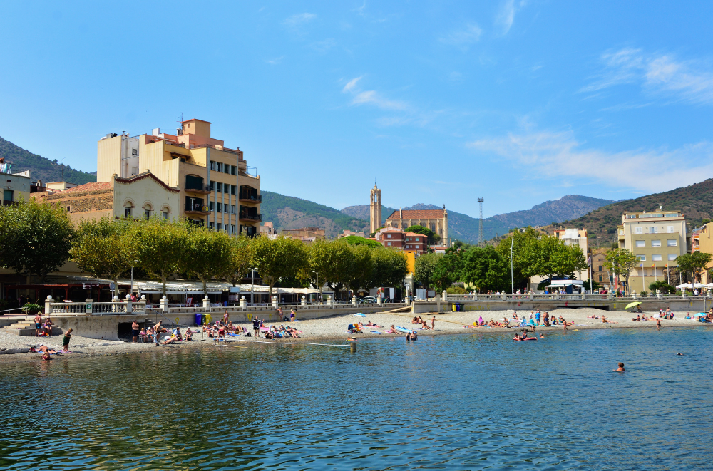 Platja de Portbou. Beachfront of a coastal town with swimmers and sunbathers, backed by colorful buildings and mountains.