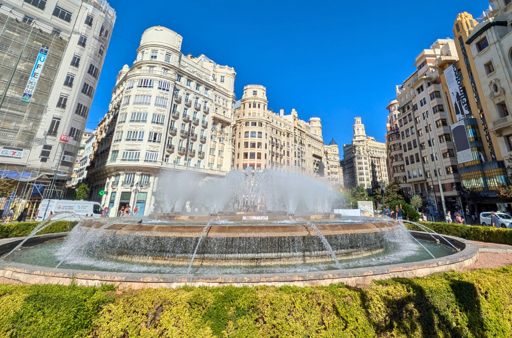 Valencia old town: Plaza del Ayuntamiento fountain with Art Nouveau buildings on a sunny day. Historic white facades and blue sky create a striking backdrop in the city center.